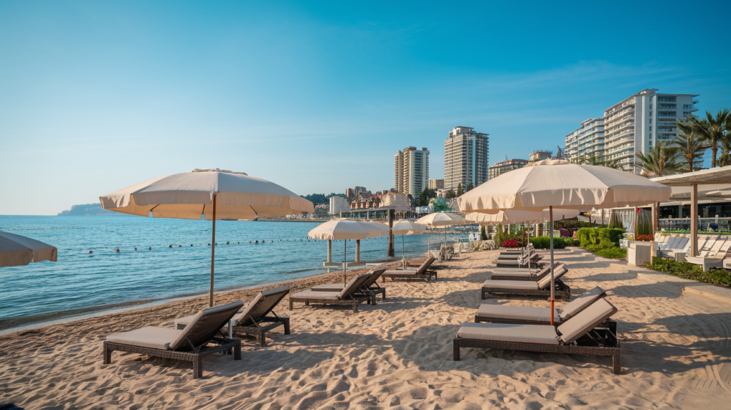 A photo of a beach club in Antalya, Turkey. There are lounge chairs and umbrellas on the sand. The background reveals a clear blue sky and towering buildings. The water is calm and the sand is golden.